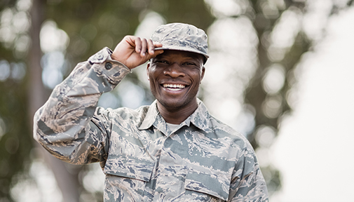 Smiling soldier in camouflage uniform tipping his hat outdoors.