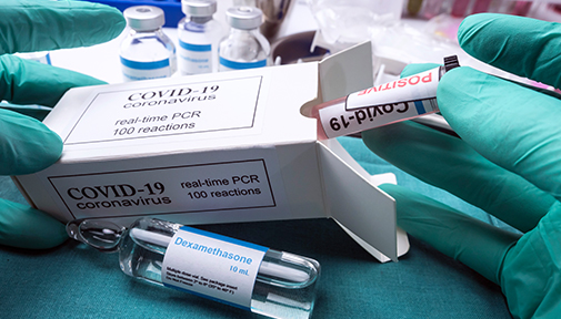 Close-up of hands in green gloves handling COVID-19 PCR test kit, a vial of dexamethasone, and other medical supplies.